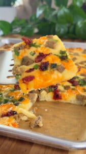 Slice of sheetpan pancake with sausage, cheese, and herbs on a spatula over a baking sheet.