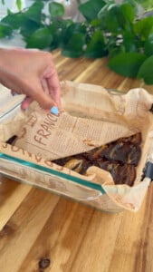 Hand lifting parchment from glass dish with dried dates on wooden table.