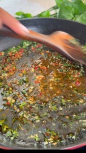 Stirring herbs and spices in sizzling oil with a wooden spoon in a black pan.