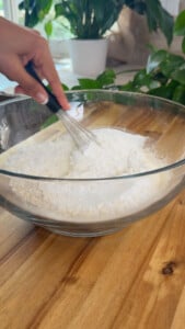 Whisking flour in a glass bowl on a wooden table, surrounded by green plants.