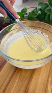 Whisking pancake batter in a clear glass bowl on a wooden countertop.