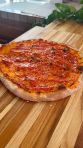 Pizza crust with tomato sauce on a wooden board in a kitchen setting.