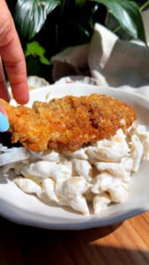 Breaded chicken fillet being placed on creamy pasta in a rustic bowl.