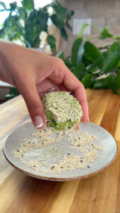 Hand holding falafel coated with sesame seeds above a plate with more seeds.