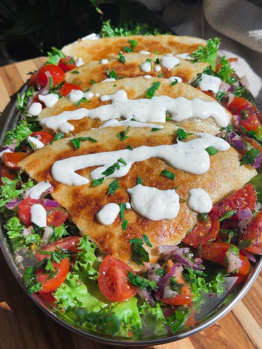 Shawarma quesadilla topped with creamy sauce, garnished with fresh tomatoes, lettuce, and herbs on a platter.