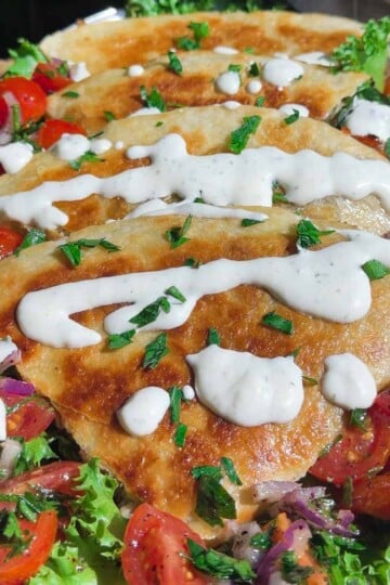 Shawarma quesadilla topped with creamy sauce, fresh tomatoes, lettuce, and herbs on a platter.