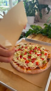 Homemade pizza with olives, artichokes, and peppers on a baking tray being covered prior to flash freeze.