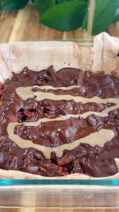 Melted chocolate and tahini over nuts and dates in a parchment-lined glass dish.