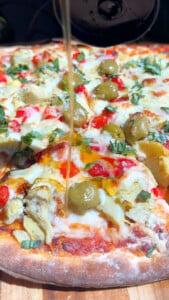 Delicious veggie pizza with olives, artichokes, and fresh herbs drizzled with hot honey on a wooden board.