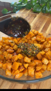Diced roasted potatoes with herb dressing in a glass bowl on wooden table, fresh green leaves in the background.