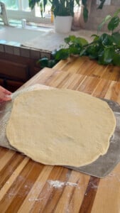 Pizza dough being shaped on a wooden counter with a kitchen background.