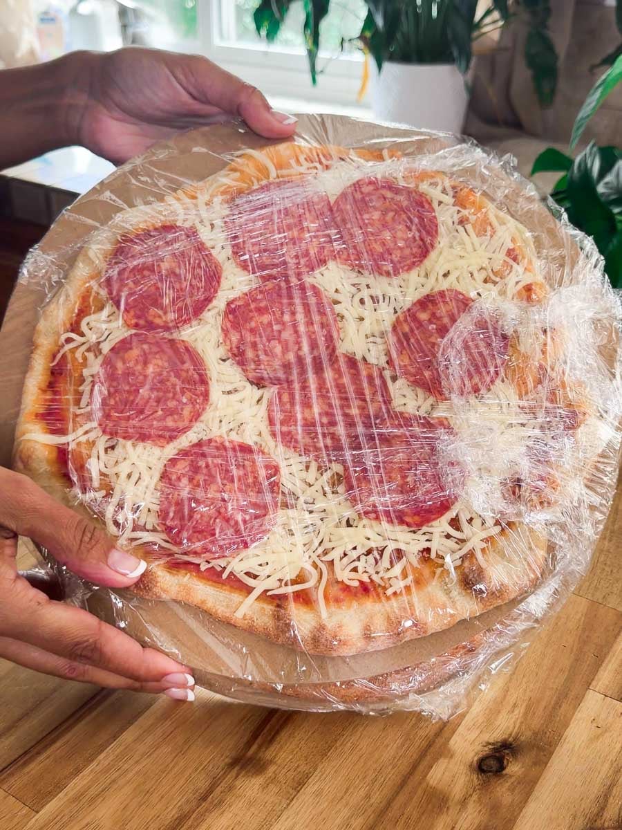 Plastic-wrapped homemade freezer pizza with cheese on a wooden surface, ready for baking.