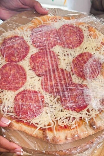 Homemade freezer pizza wrapped in clear plastic on a wooden table.
