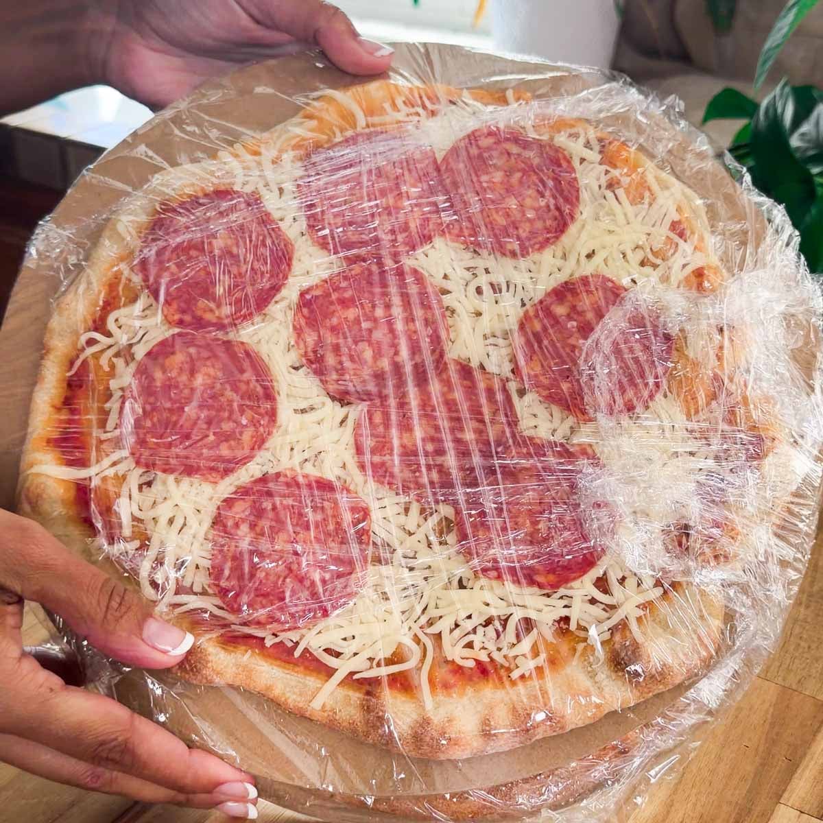 Homemade freezer pizza wrapped in clear plastic on a wooden table.