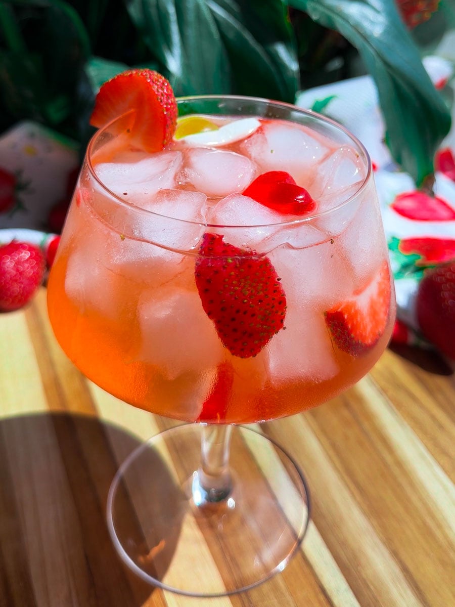 Refreshing Homemade Strawberry Gin cocktail with ice in a large glass on a wooden table, garnished with fresh strawberries and cherries.
