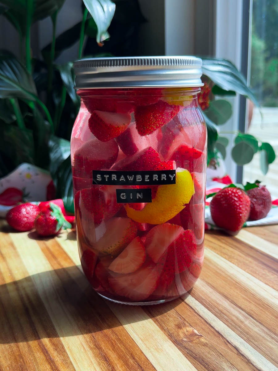 Jar of homemade strawberry gin with lemon on wooden table surrounded by fresh strawberries.