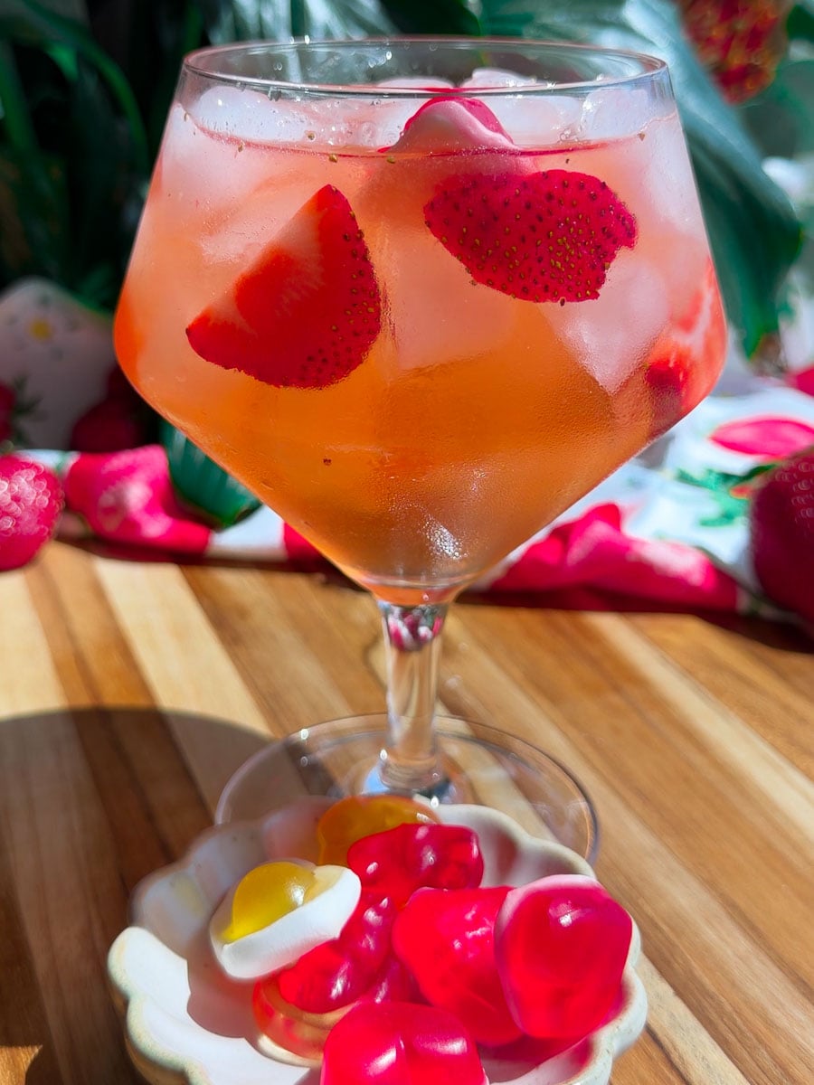 Refreshing Homemade Strawberry Gin cocktail with ice, paired with a dish of colorful gummy candies on a wooden table.