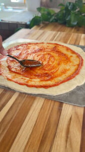 Spreading tomato sauce on pizza dough with a spoon on a wooden countertop.