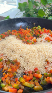Rice cooking with colorful diced vegetables in a skillet, surrounded by greenery.