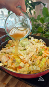 Pouring broth over cabbage in a skillet on a wooden countertop, with green plants in the background.