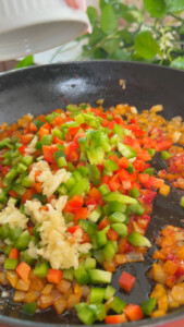 Chopped peppers and garlic in a frying pan for sautéing with greenery in the background.