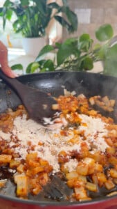 Cooking onions and spices with flour in a pan, kitchen plants in the background.