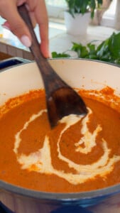 Stirring cream into tomato soup in a pot with a wooden spoon, kitchen background.