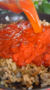Pouring tomato sauce over cooked ground meat and onions in a pan.