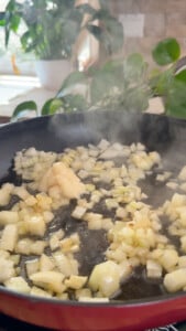 Chopped onions sautéing in a pan with cooking fat amidst kitchen plants.