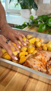Seasoned chicken and potatoes in a baking tray on wooden surface, hands arranging ingredients.