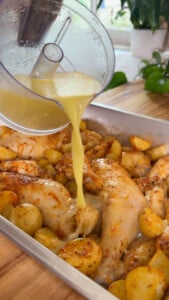 Pouring garlic lemon sauce over seasoned chicken and potatoes in a baking tray.