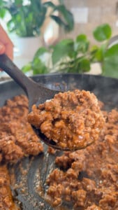 Cooking ground beef in a skillet with a spoon, fresh ingredients in the background.