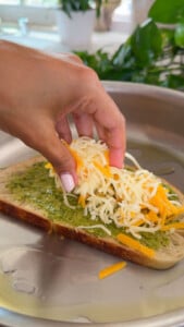 Hand adding shredded cheese to pesto-covered bread in a pan for making a grilled cheese sandwich.