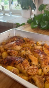 Oven-baked chicken and potatoes in a roasting pan by a kitchen window with green plants.