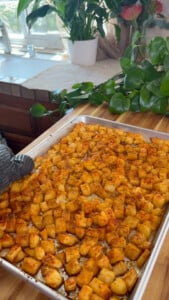 Roasted seasoned potatoes on a baking tray, surrounded by lush green plants in a bright kitchen setting.