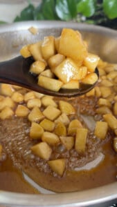 Cooking diced apples in cinnamon syrup on a wooden spoon over a pot.