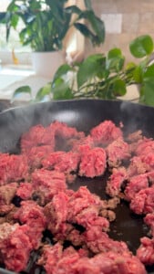 Ground beef cooking in a skillet, with steam rising and plants in the background.