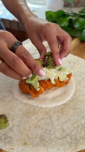 Person preparing wrap with crispy chicken, falafel, lettuce, and sauce on a tortilla.