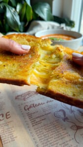 Cheesy grilled cheese sandwich split open, with tomato soup in the background.