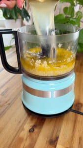 Pouring liquid into a blue food processor with chopped garlic and oil on a wooden countertop.