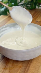 Creamy white sauce being poured from spoon into a bowl on a wooden table.