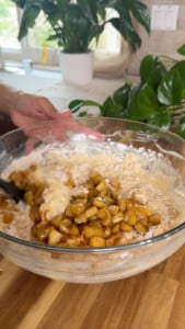 Mixing apple pieces into batter in a glass bowl on a wooden countertop.