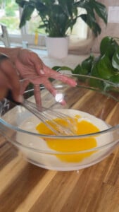 Hand whisking eggs and milk in a clear bowl on a wooden countertop.