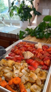 Tray of roasted vegetables with tomatoes, onions, carrots, and garlic, in a kitchen setting with plants.
