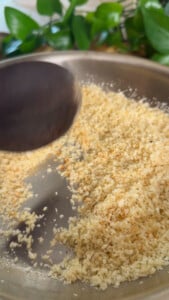 Toasting breadcrumbs in a skillet with a wooden spoon, green leaves in the background.