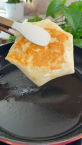 Golden-brown stuffed crunchy wrap cooking on skillet with spatula, surrounded by green plants in a bright kitchen.