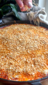 Hand sprinkling breadcrumbs on baked dish with golden crust in black skillet.