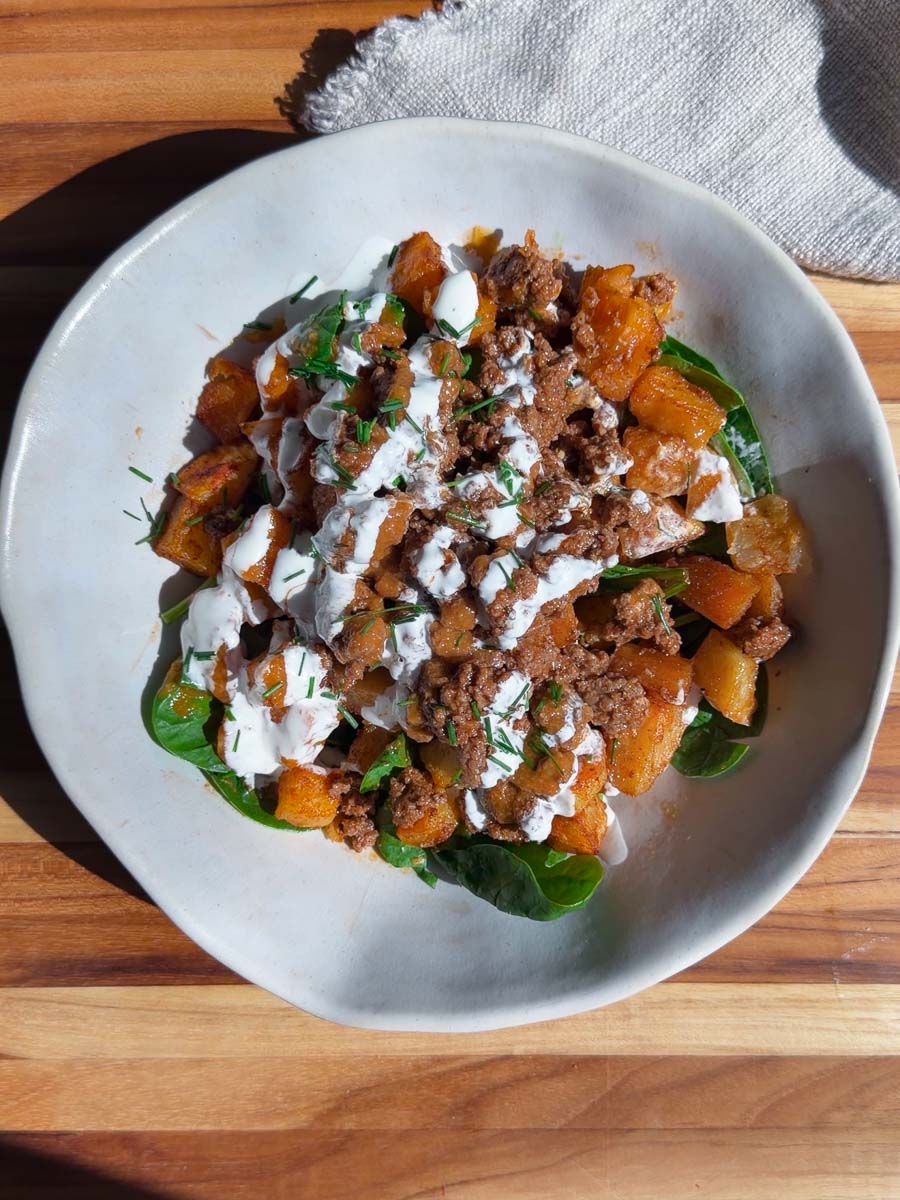 Patata brava bowl with ground meat, roasted squash, fresh greens, and cream sauce on a wooden table.