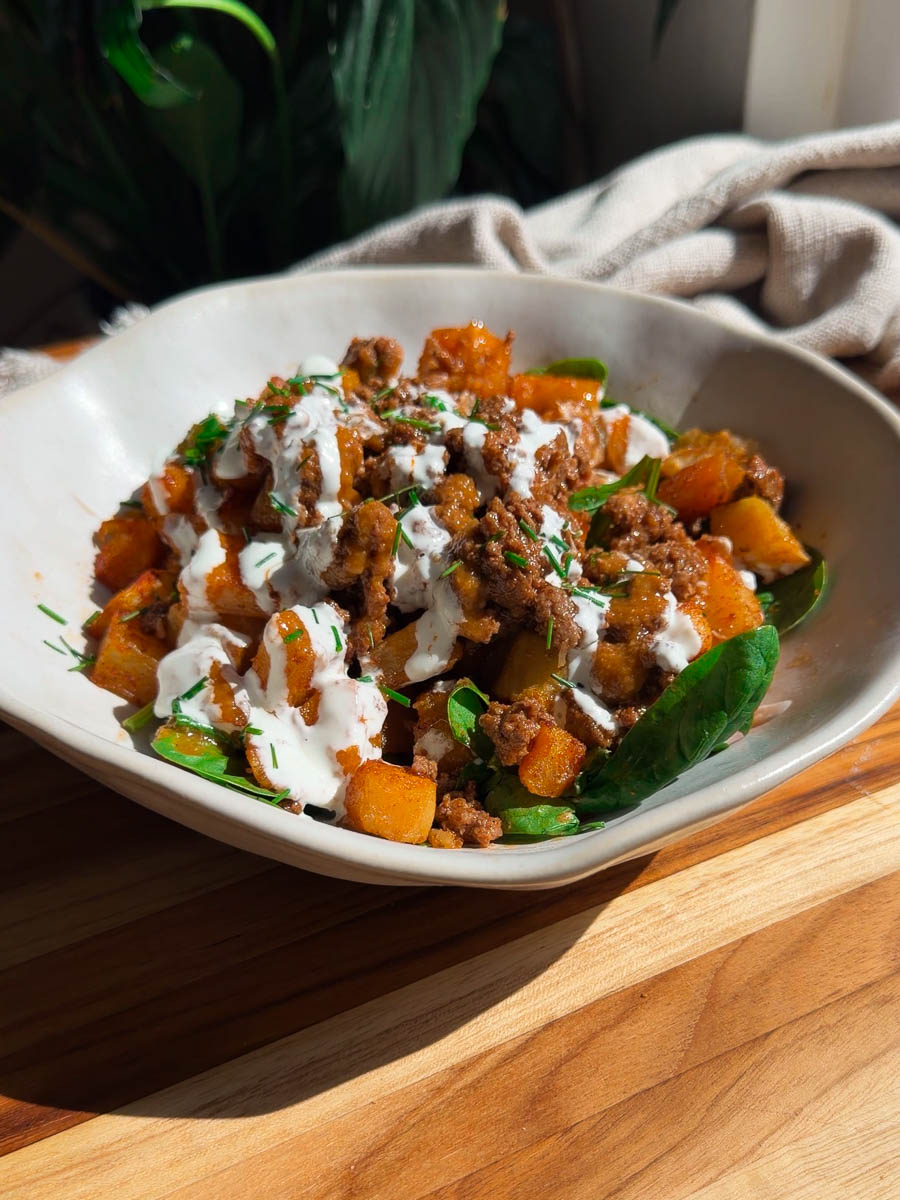 Patata brava bowl with ground meat, fresh greens, and drizzled yogurt sauce on a wooden table.