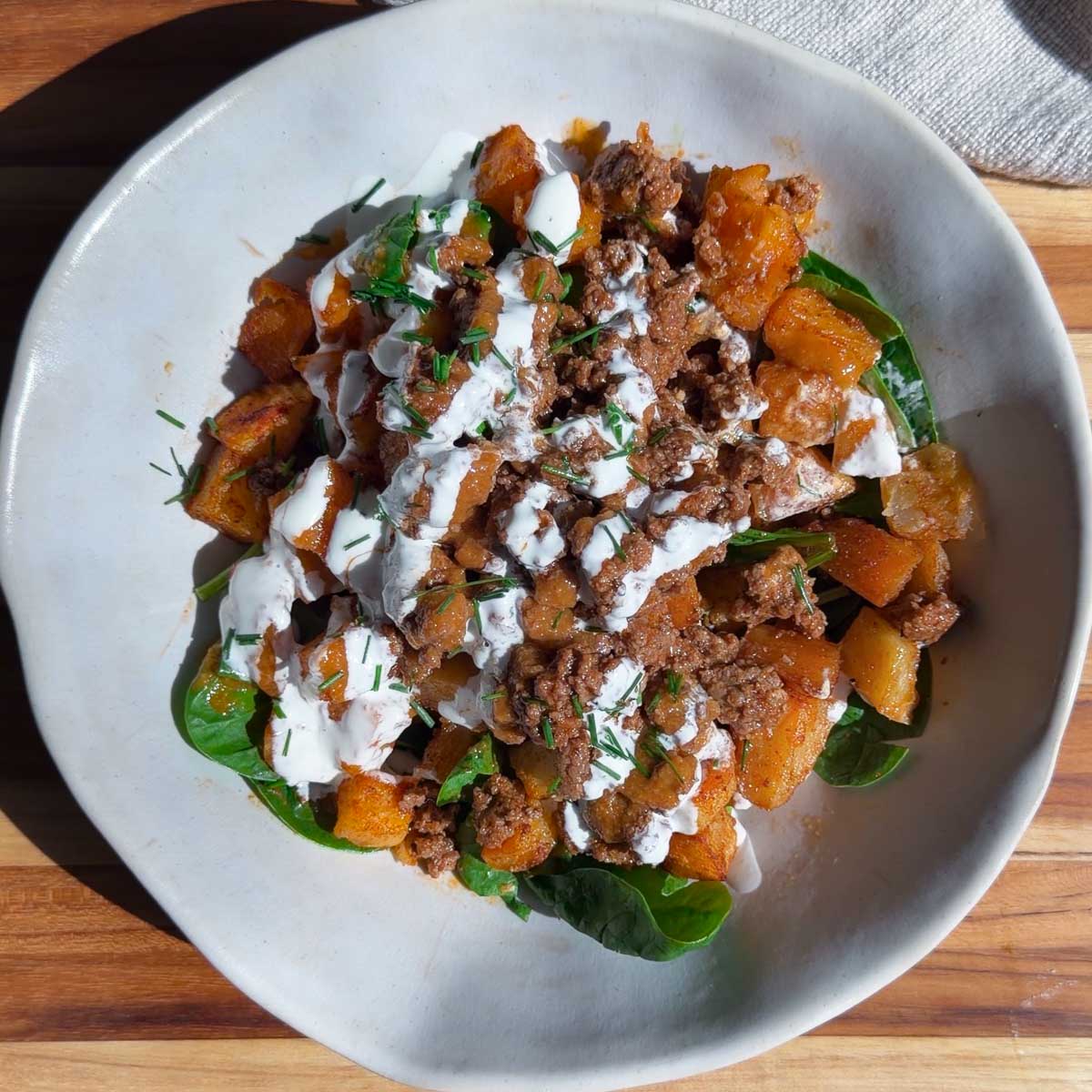 Patata brava bowl with ground beef with herbs and creamy drizzle on a wooden table.